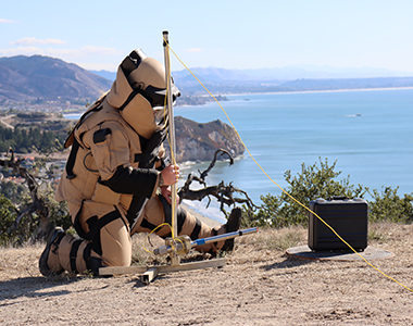 Bomb Task Force member wearing a bomb suit and using a water disruptor on an explosive briefcase with the ocean in the background.