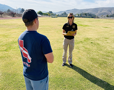 Backgrounds manager smiling holding a clipboard with a man in a t-shirt and jeans with his back to the photo looking at her standing in a field.