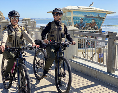 Two bicycle patrol team members standing with their bikes on a promenade with the ocean in the background.