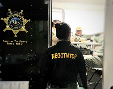 Crisis Negotiation Team (CNT) Sergeant's back showing a jacket with the word "Negotiator" with a Sheriff's Office trailer in front of her with the door open showing blurred Special Enforcement Team members.