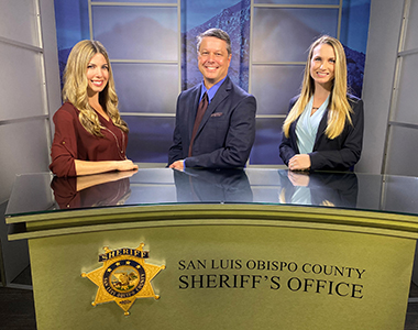 Three Crime Prevention Unit members standing behind a media desk looking forward smiling.