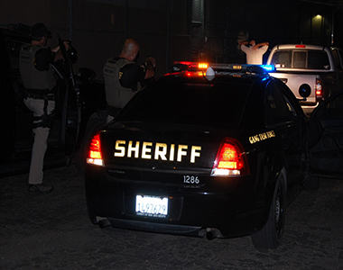 Two Gang Task Force members standing next to their vehicles with guns pointed at a subject in front of them standing outside of a white truck.