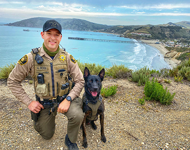 Sheriff's Office K9 Handler kneeling with K9 Mando with Avila Beach in the background.