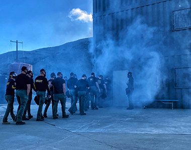 Mutual Aid and Public Safety (MAPS) team members enter a building wearing gas masks with gas coming out of the door.