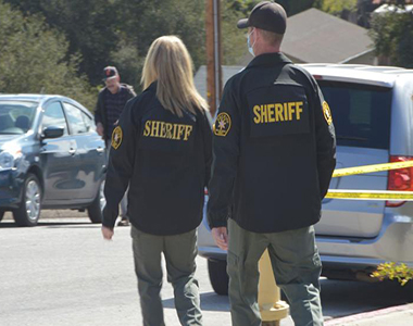 Two Major Crimes detectives walking away with crime scene tape to their right and two vehicles beyond them.