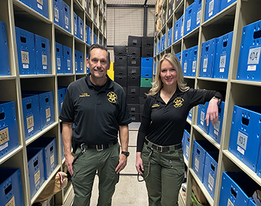 Two Property Officers standing in a hallway with blue property boxes on both sides on shelves.