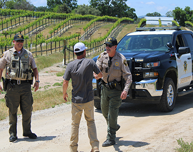 Rural Crime Unit Deputies shaking hands with a rancher with vineyards in the background and a patrol truck.